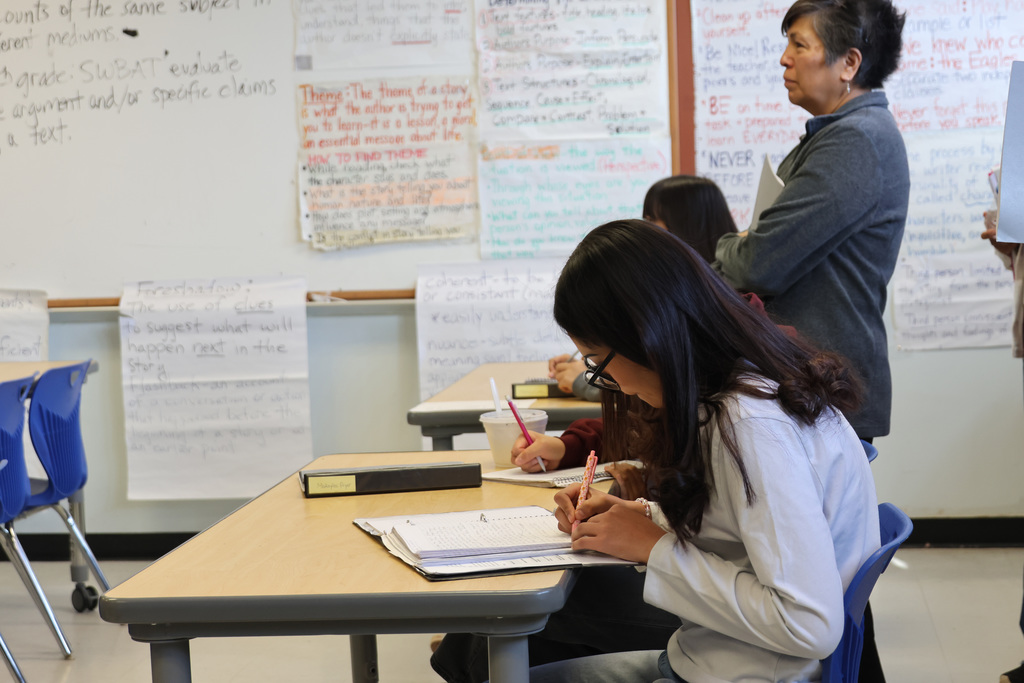 A classroom with a student writing in a notebook. A teacher stands by, and various papers are on the wall.