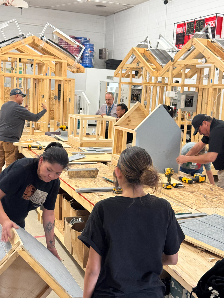 Workers in a workshop, constructing wooden structures. One woman bent over a frame, another stands observing. Various tools on the table.