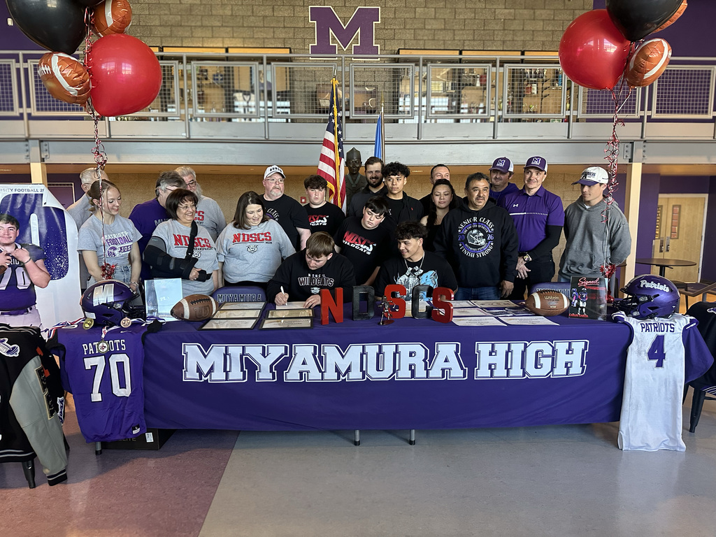 Group of people, possibly athletes, gather around a table with a banner reading "Miyamura High".