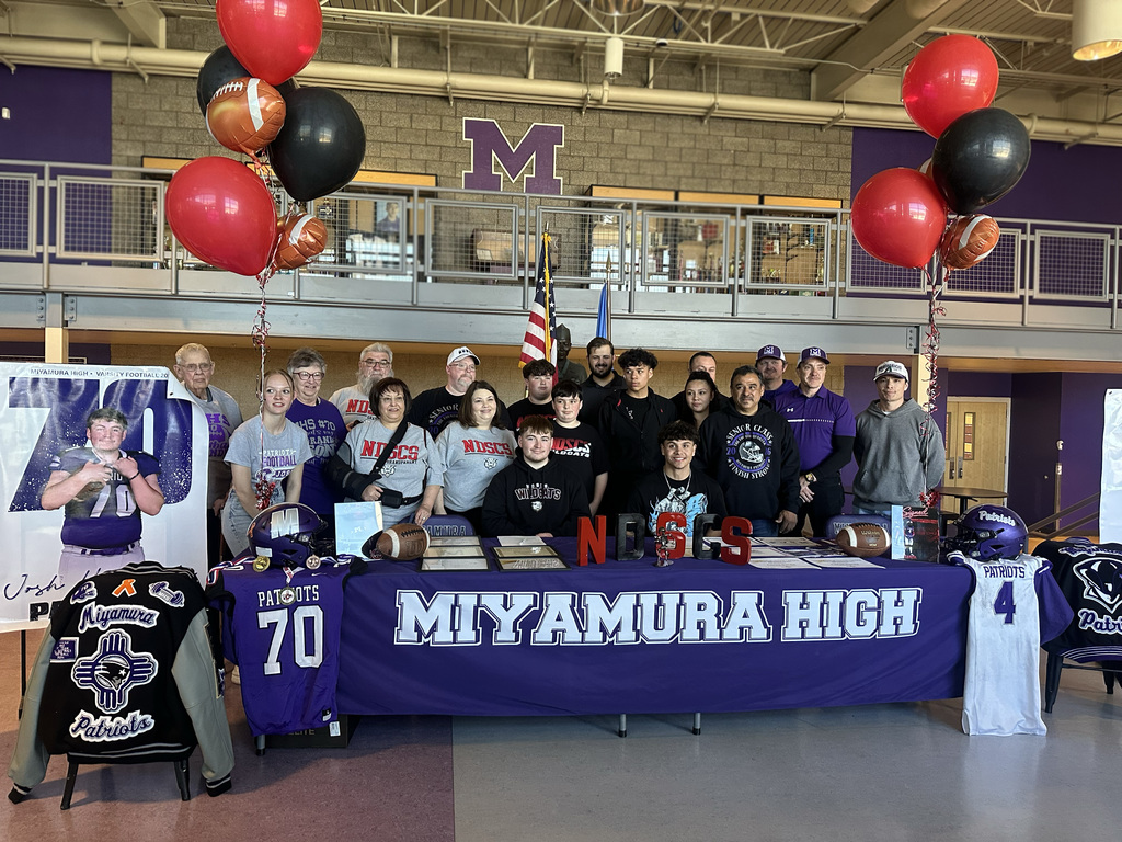 Group photo of people at a table. The table has a banner reading "Miyamura High". Sports gear and balloons are in the background.