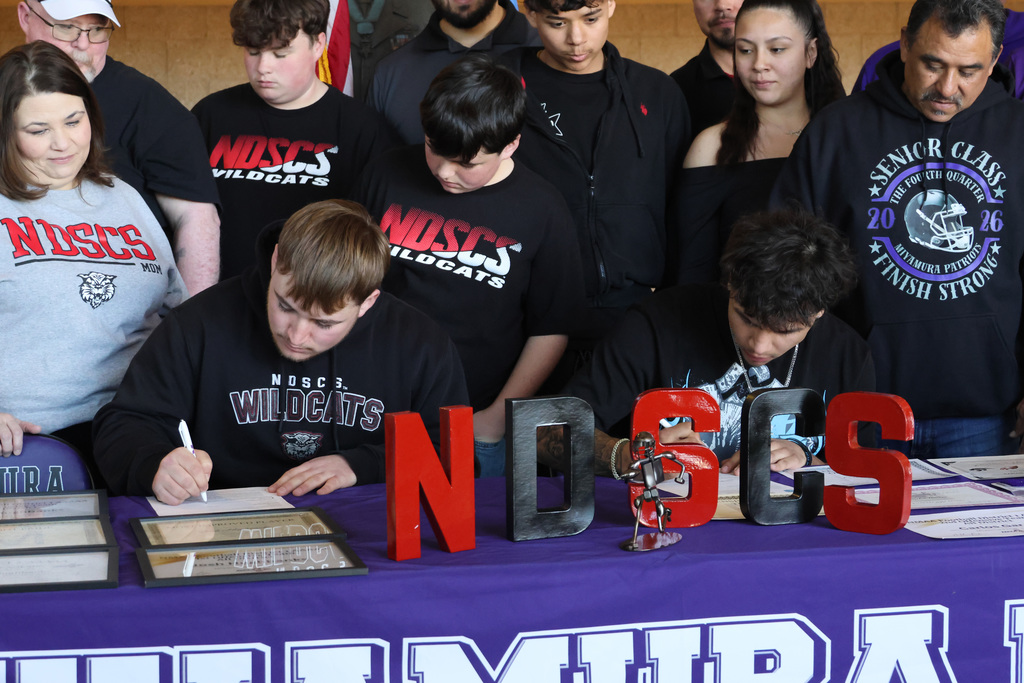 People in team jerseys, some signing documents on a table with letters "NDHS" and "UNMIDA" on a purple cloth.