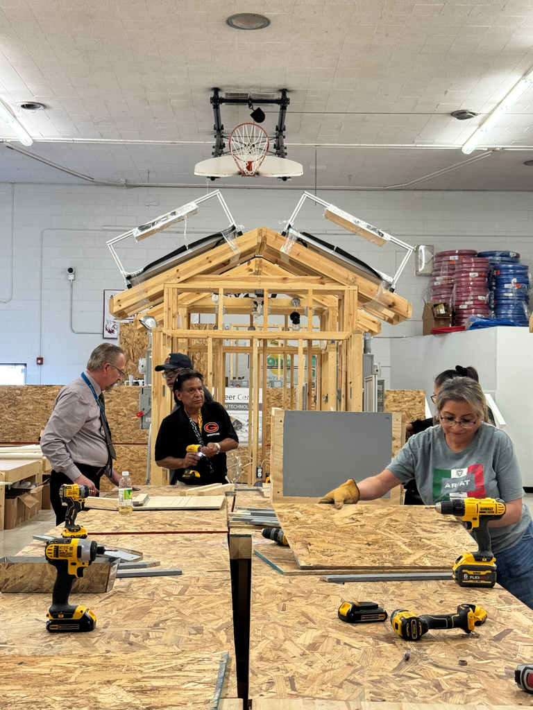A group of people work in a workshop on wooden planks, using drills and tools. A house model is under construction in the background.