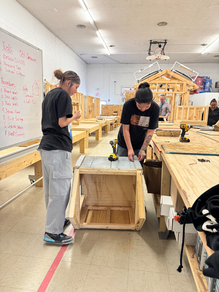 Two people work on a wooden box at a workbench in a workshop. A whiteboard is on the wall behind them.