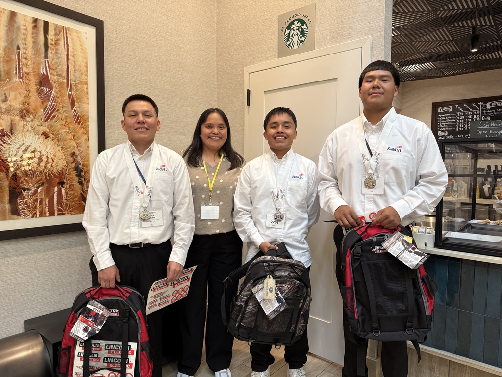 Four people stand in a room with bags. One holds a red bag. Another holds a black bag. One wears a necklace.