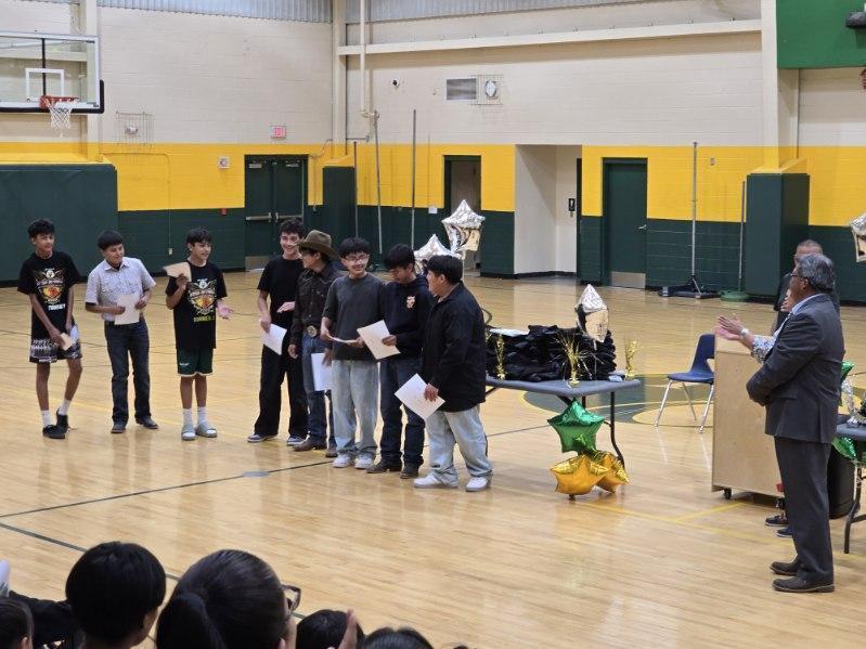 Several people stand on a basketball court holding papers, one speaking at a podium. Audience sits on chairs.