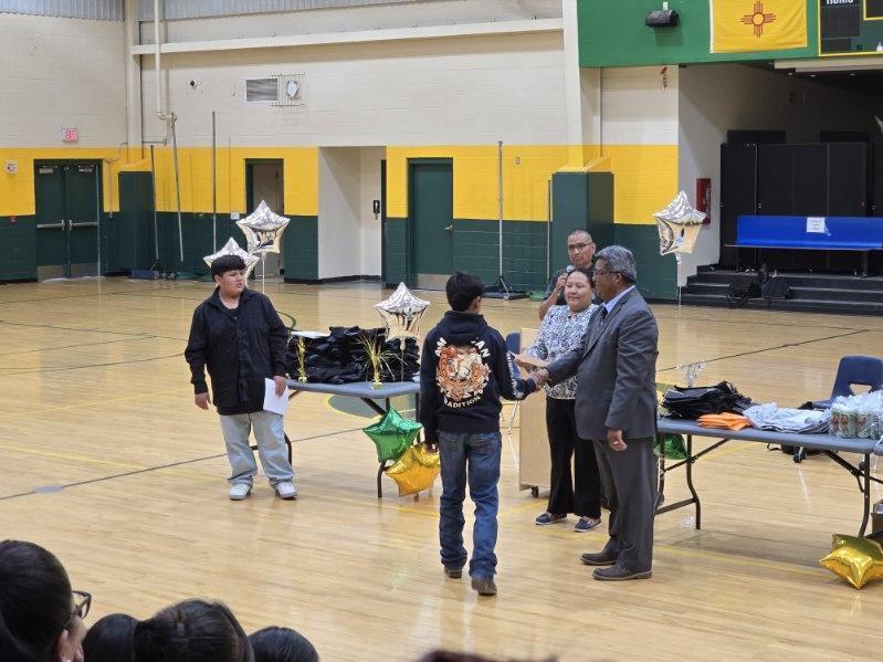 An indoor basketball court hosts an event with people standing around tables, including two individuals shaking hands.