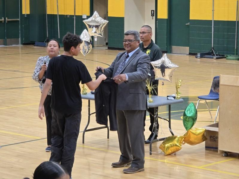 A man in a suit hands a jacket to a boy. They are in a gym with a blue and yellow wall.