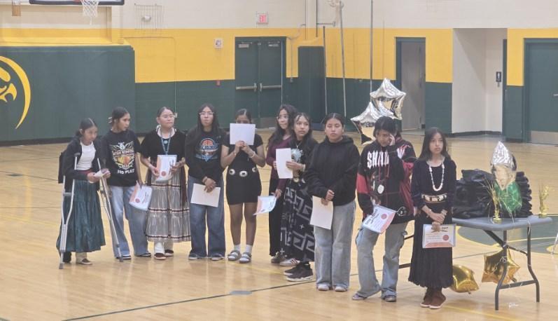 Eleven individuals, dressed in various outfits, hold papers in a gymnasium. They stand in front of a table with items.