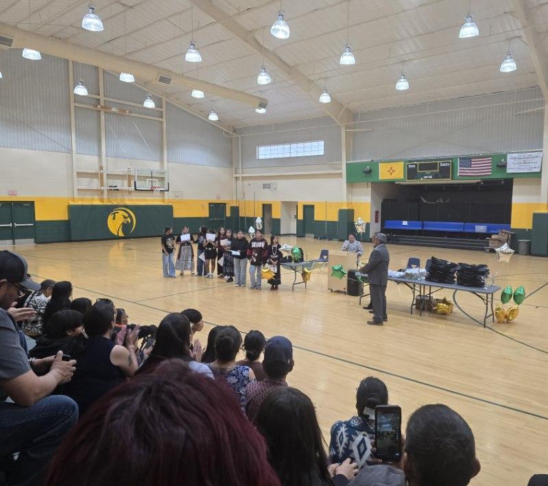 A group of people stand on a basketball court as a person speaks at a table. Audience members watch from the bleachers.