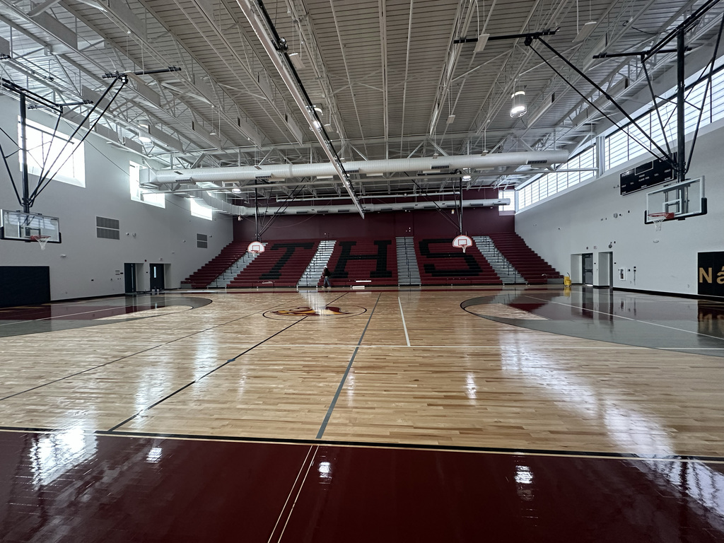 Indoor basketball court with hardwood flooring and white and red bleachers. Large windows and bright lighting.