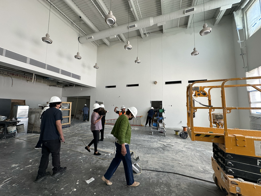 A group of people in white hard hats walk in a large, unfinished room with exposed pipes.