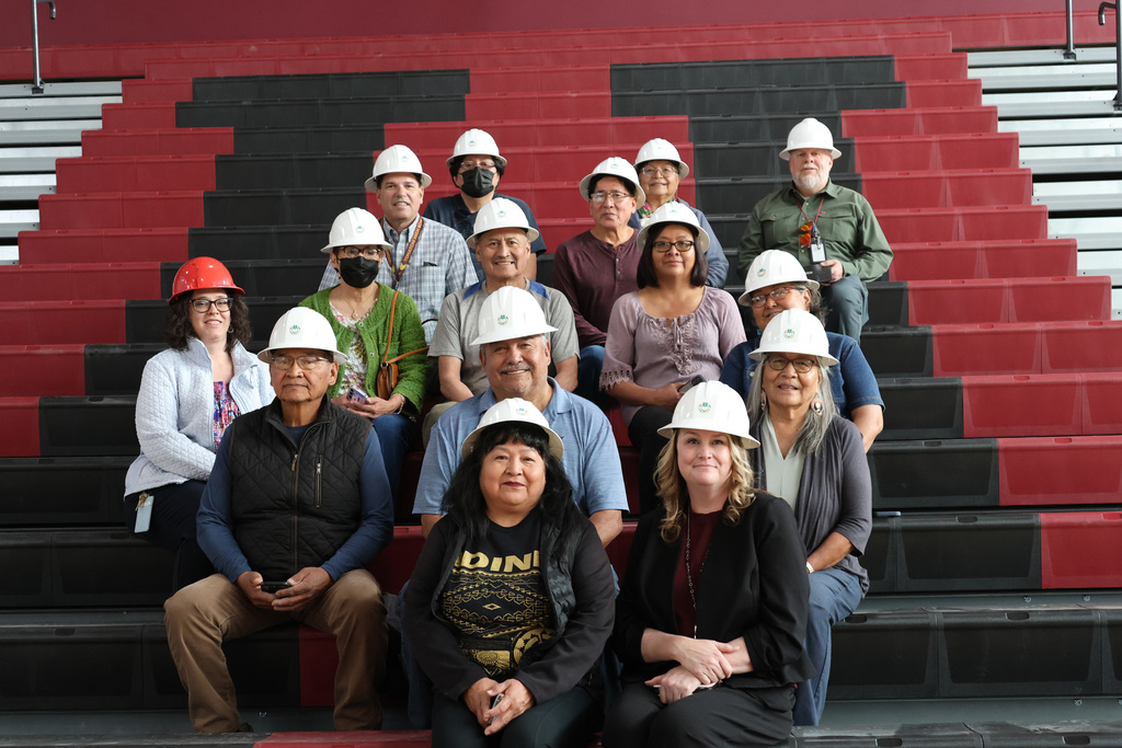 A group of individuals, all wearing white hard hats, seated on a red bleacher. Some wear face masks.
