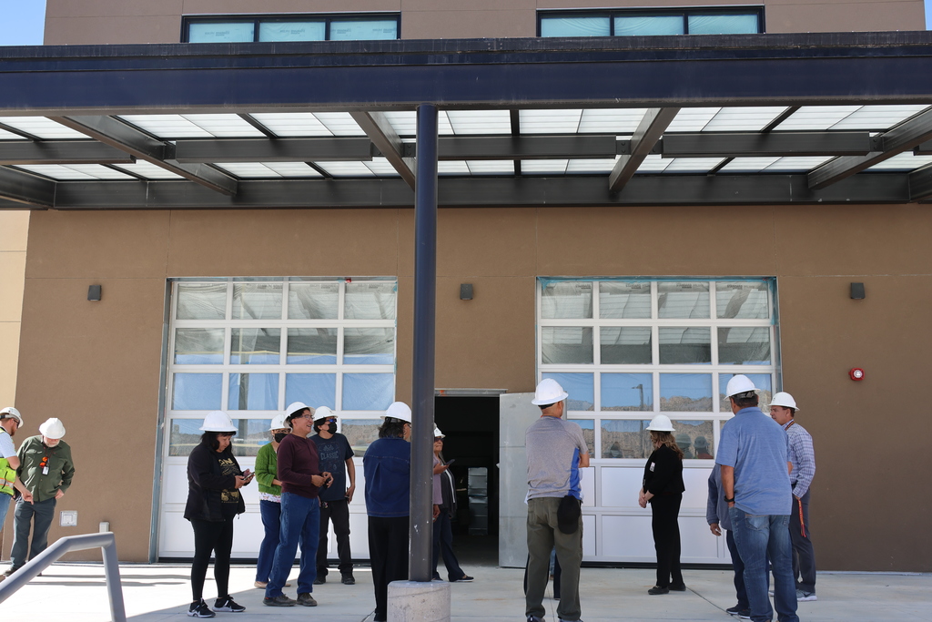 People wearing white hard hats are standing outside a tan building with large glass doors.