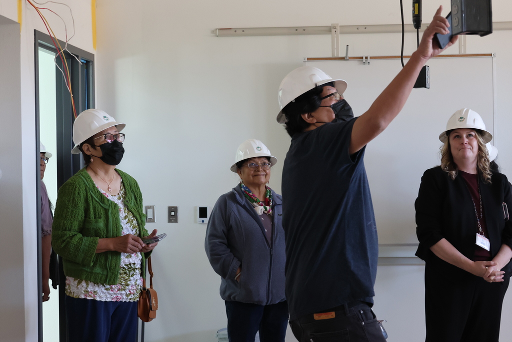 Four individuals in a room, all wearing white hard hats. One man in a blue shirt points upward.