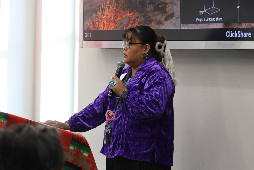 A woman in a purple shirt speaks into a microphone in front of a screen. A red cloth is on the table.