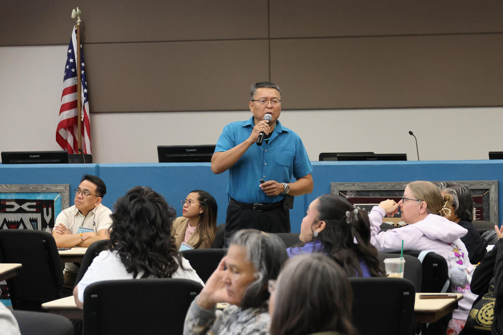 a man holds a microphone and talks to an audience. he is wearing a blue shirts, black pants, he has a watch on his left writst