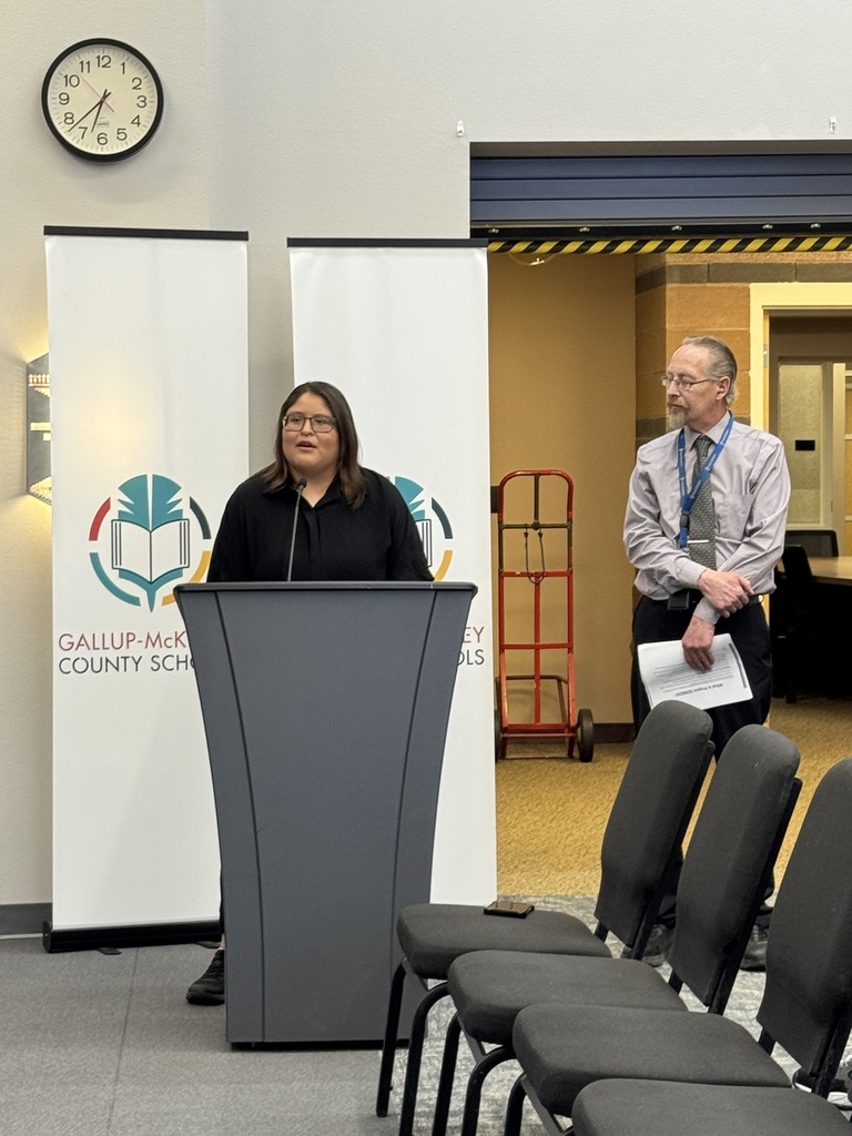 A woman speaks at a podium in front of an audience. Banners with logos are behind her.