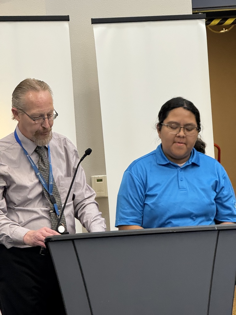 Two people stand behind a podium with microphones; one wears a blue shirt, the other a gray shirt with a tie.