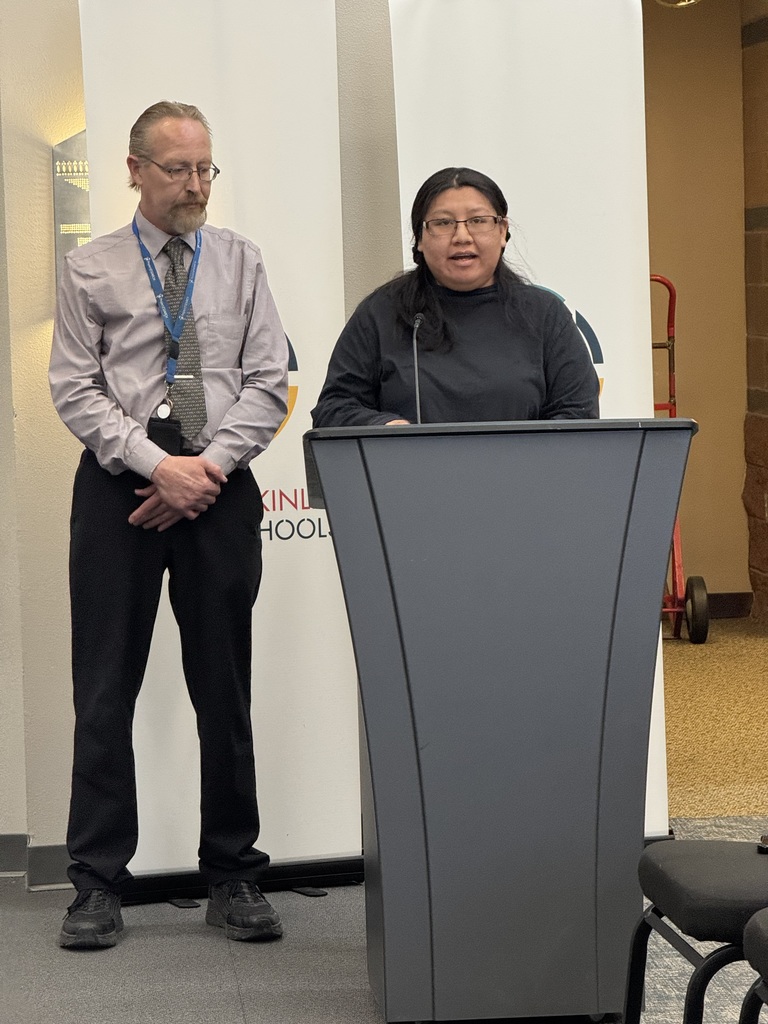 A person with glasses speaks at a podium, another person in glasses and a tie stands behind them.