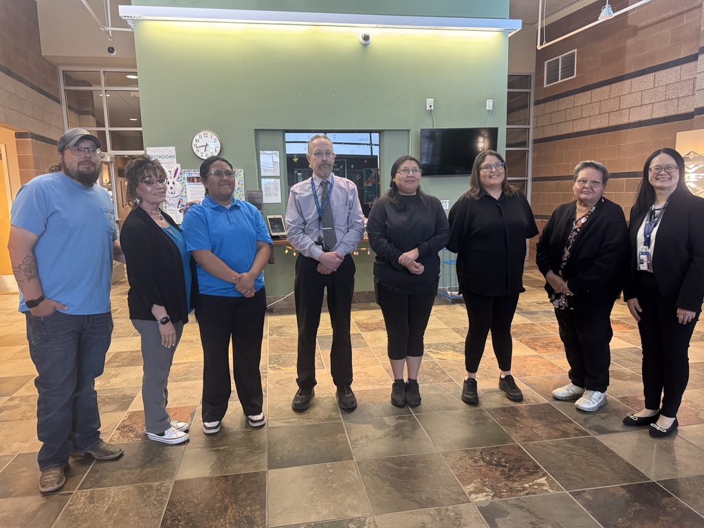 A group of seven people stand in a tiled room with a green wall and clock, likely posing for a photo.