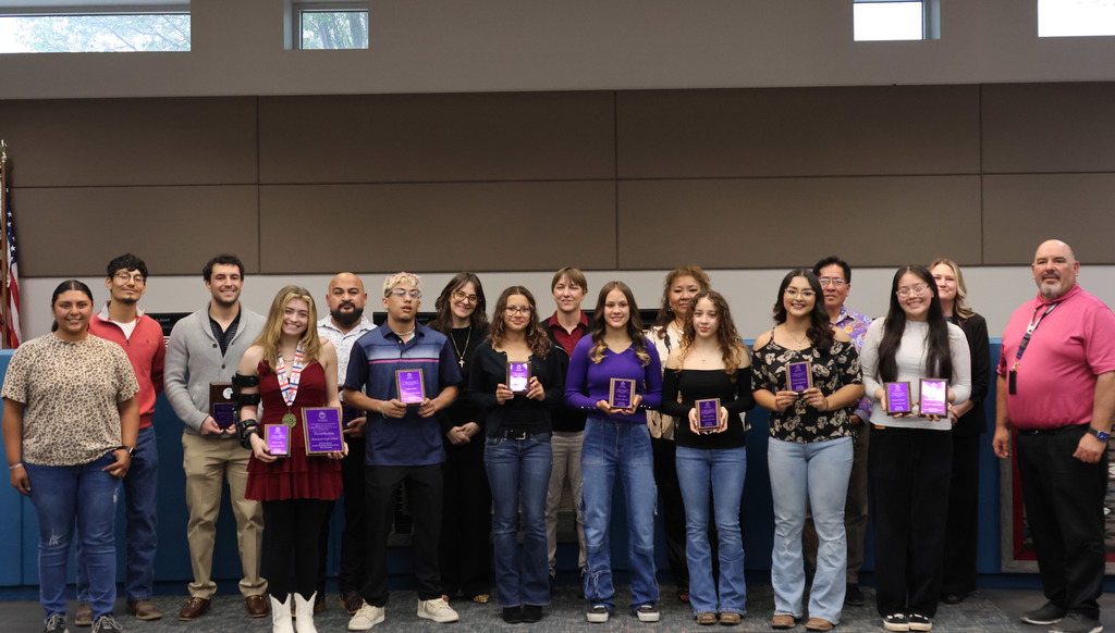 A group of people stand in a room, holding awards. They are dressed in casual clothing.