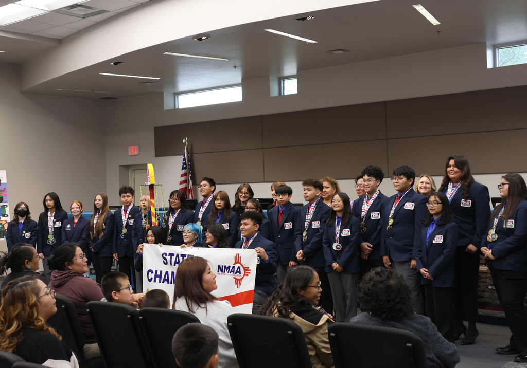 A group of people in blue suits, some holding a sign, stand in a room with chairs and spectators.