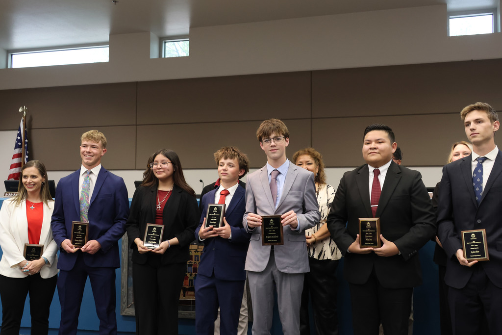 A group of individuals in suits and ties, holding awards, standing in a room with an American flag.