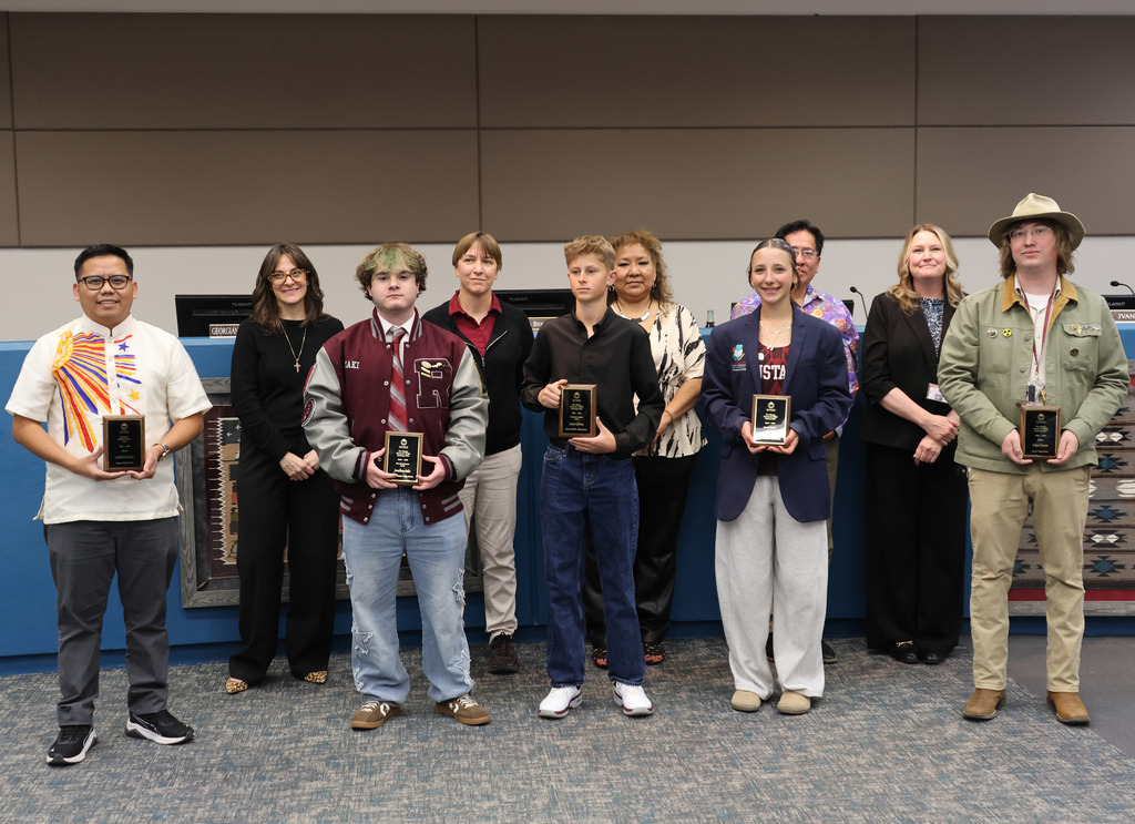 A group of people stand behind a desk, holding awards. Some wear formal attire, others casual. Two have hats. The wall is light blue.