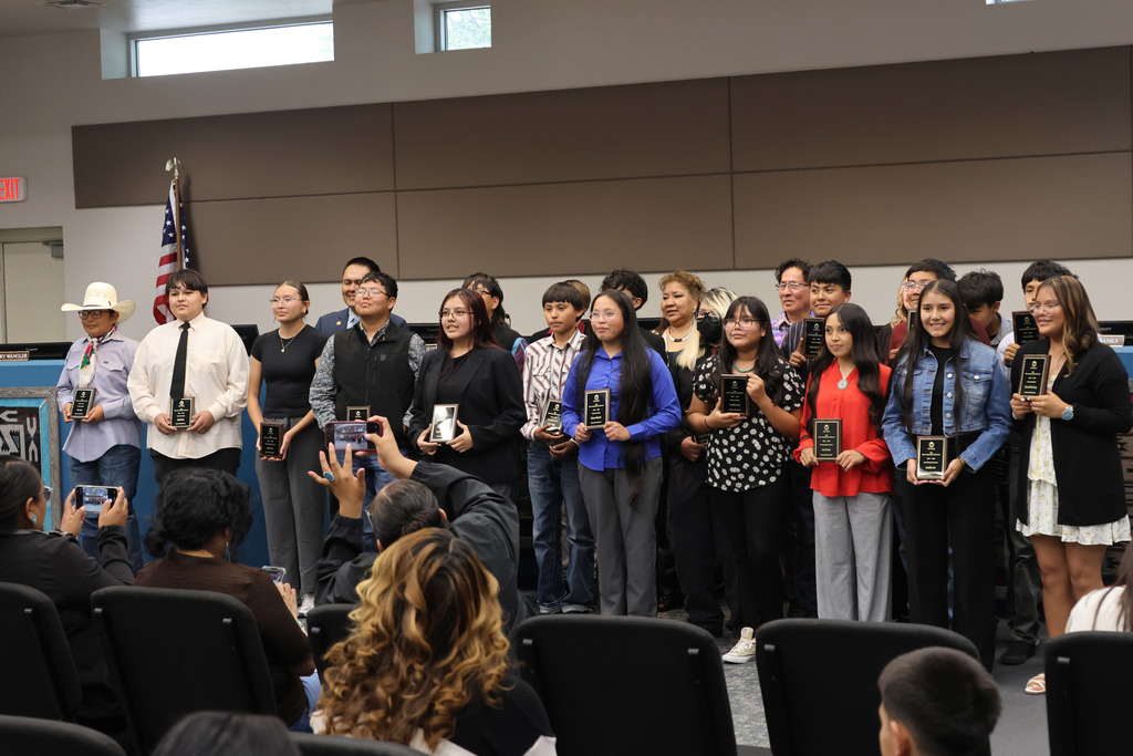 A group of people stands on a stage, holding awards, in a room with rows of chairs.