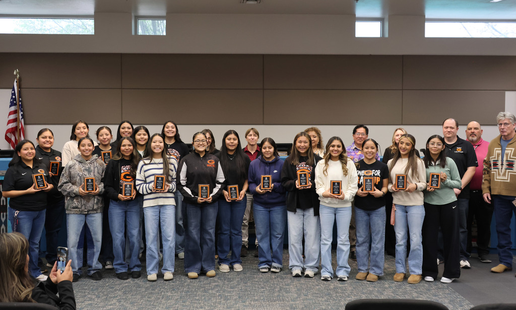A group of people in jeans and jackets pose with awards in a room with a flag.