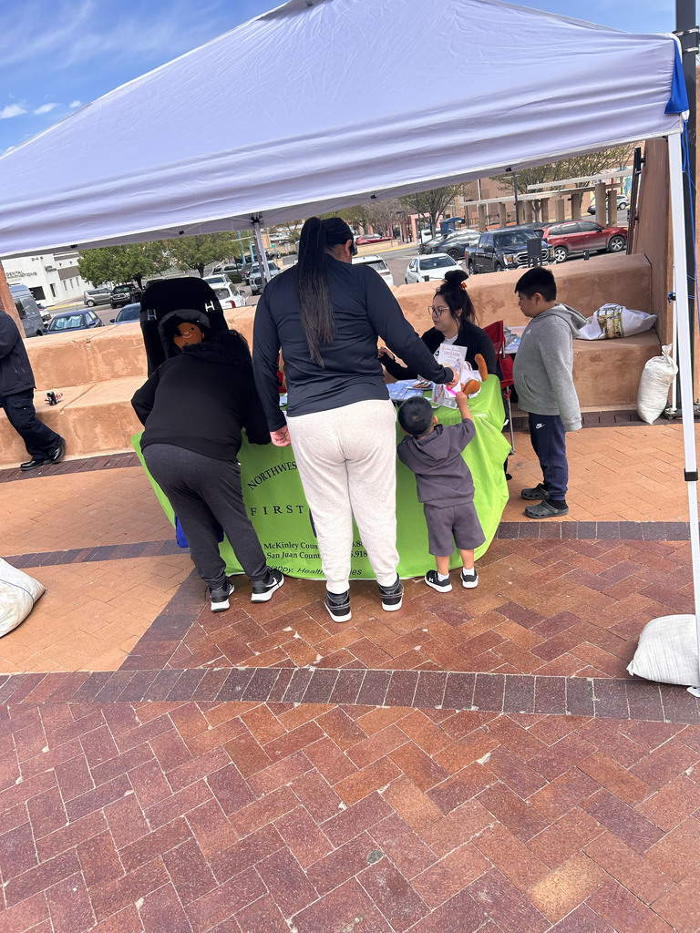A group of people stand around a green table under a canopy. A woman assists a child standing on the table.