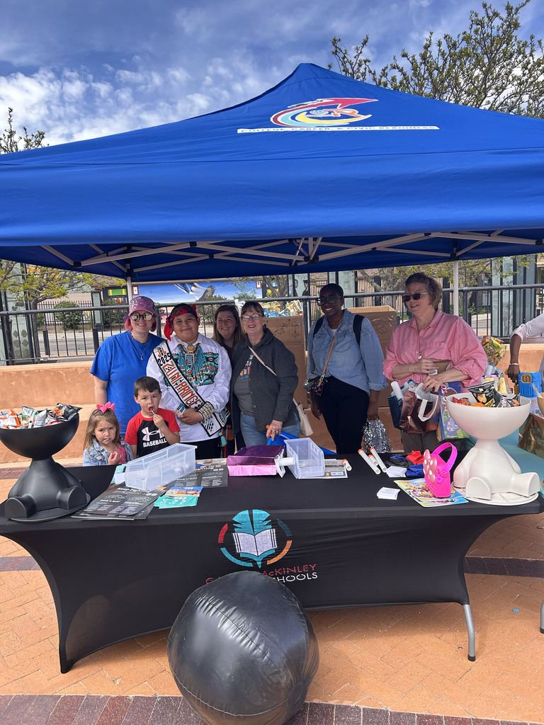 People stand under a blue canopy near a table with books and bags. The table is in front of a brick wall.