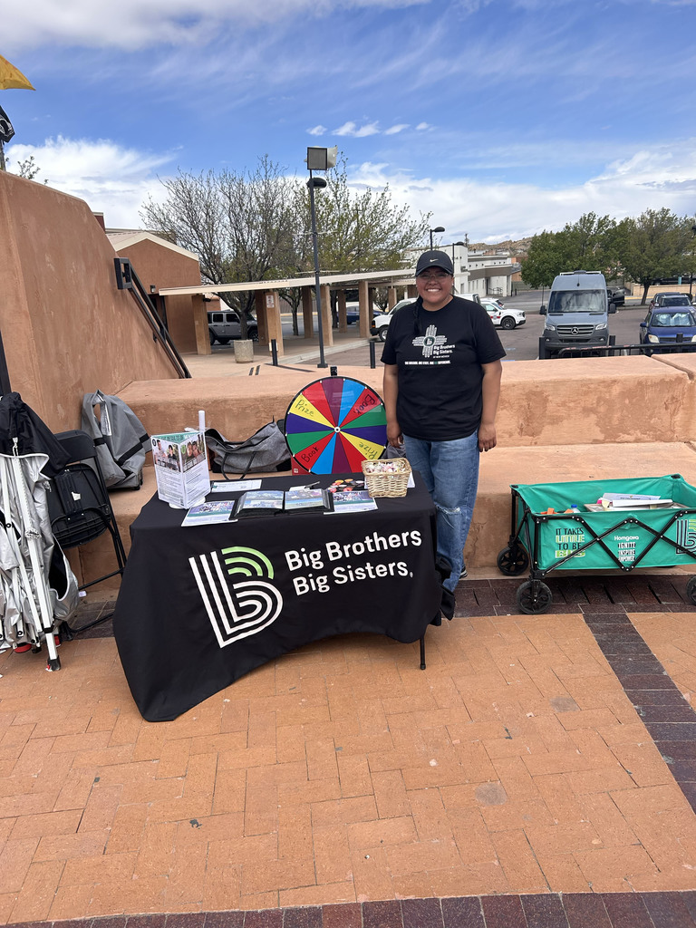A person stands at a table covered with a black cloth and a logo. Behind them, a colorful wheel sits on the table.