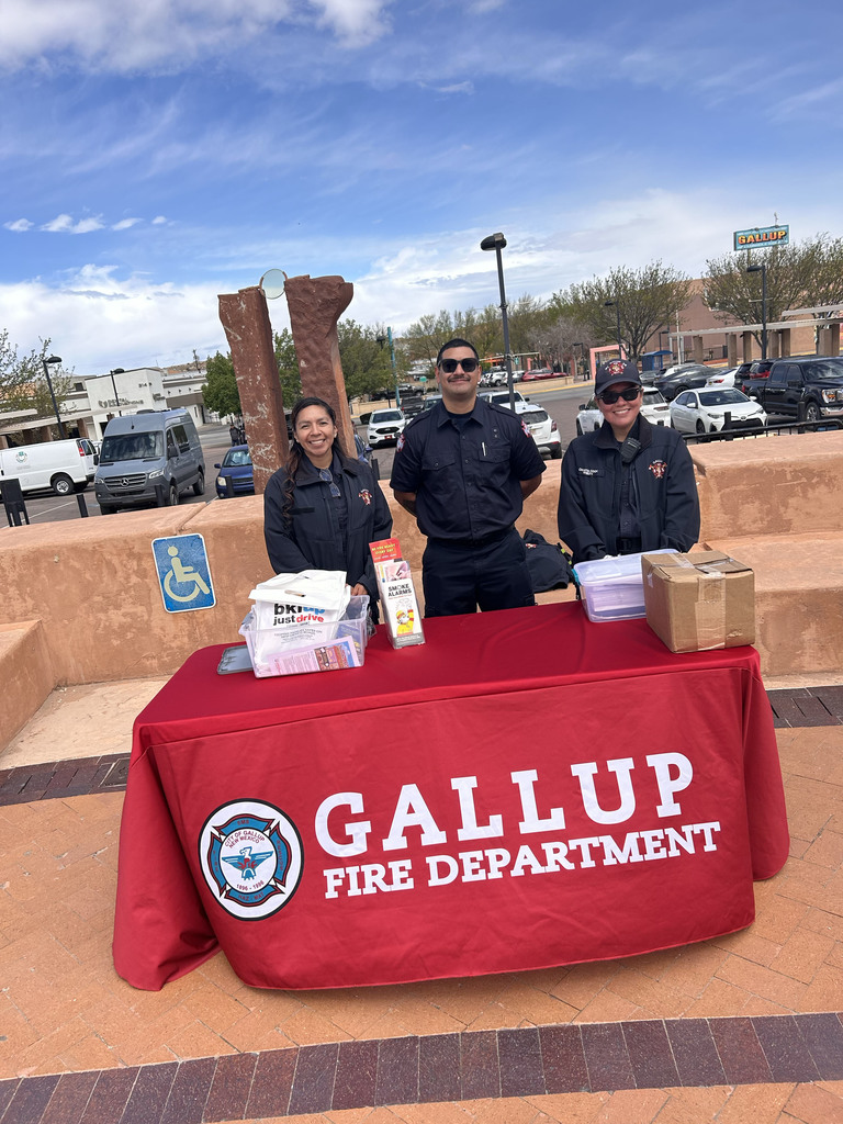 Three people stand behind a table with a red cloth that says Gallup Fire Department. They wear uniforms. Behind them, a stone wall and parked cars are visible.