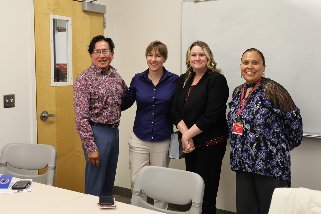 Five people stand in a room, smiling, with two chairs behind them and a table in front.