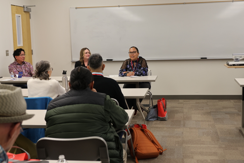 Four people sit at a table in a classroom. One person speaks into a microphone. Other people sit in chairs facing them.