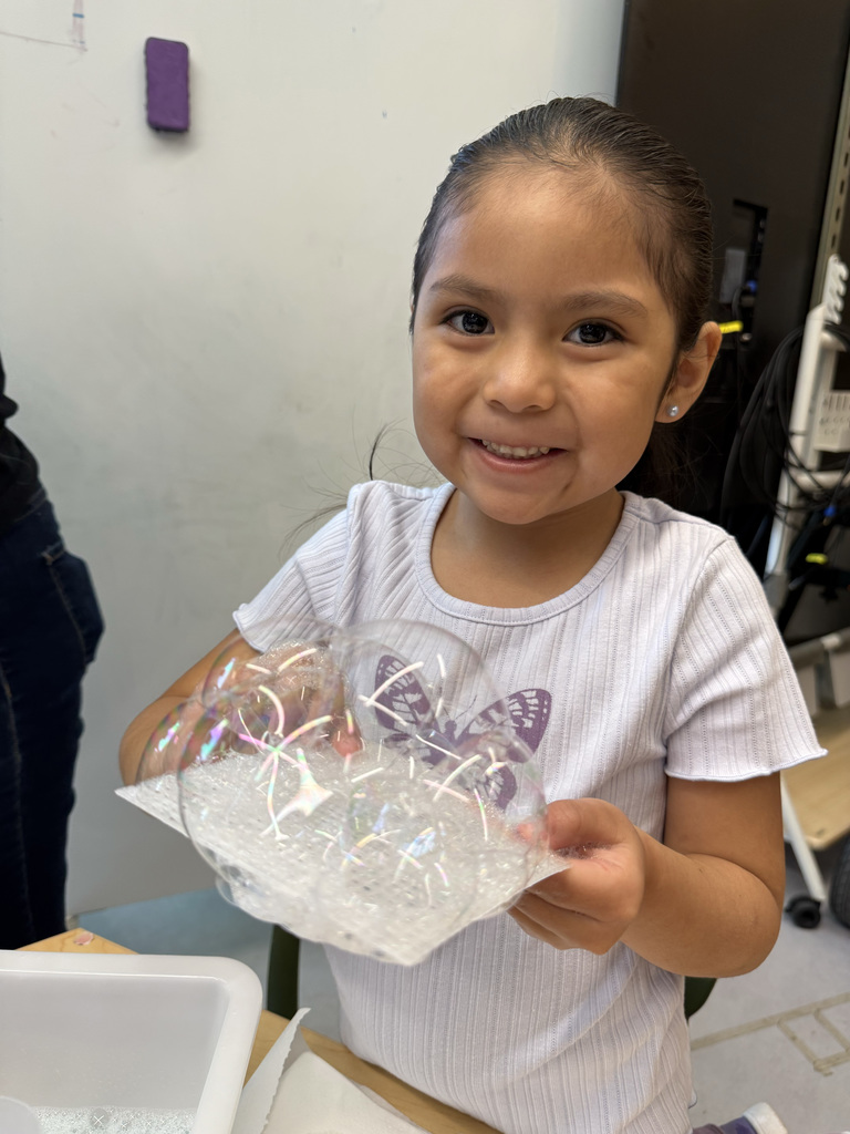 Girl holding a plastic bubble wand, smiling, standing near a table and white walls.