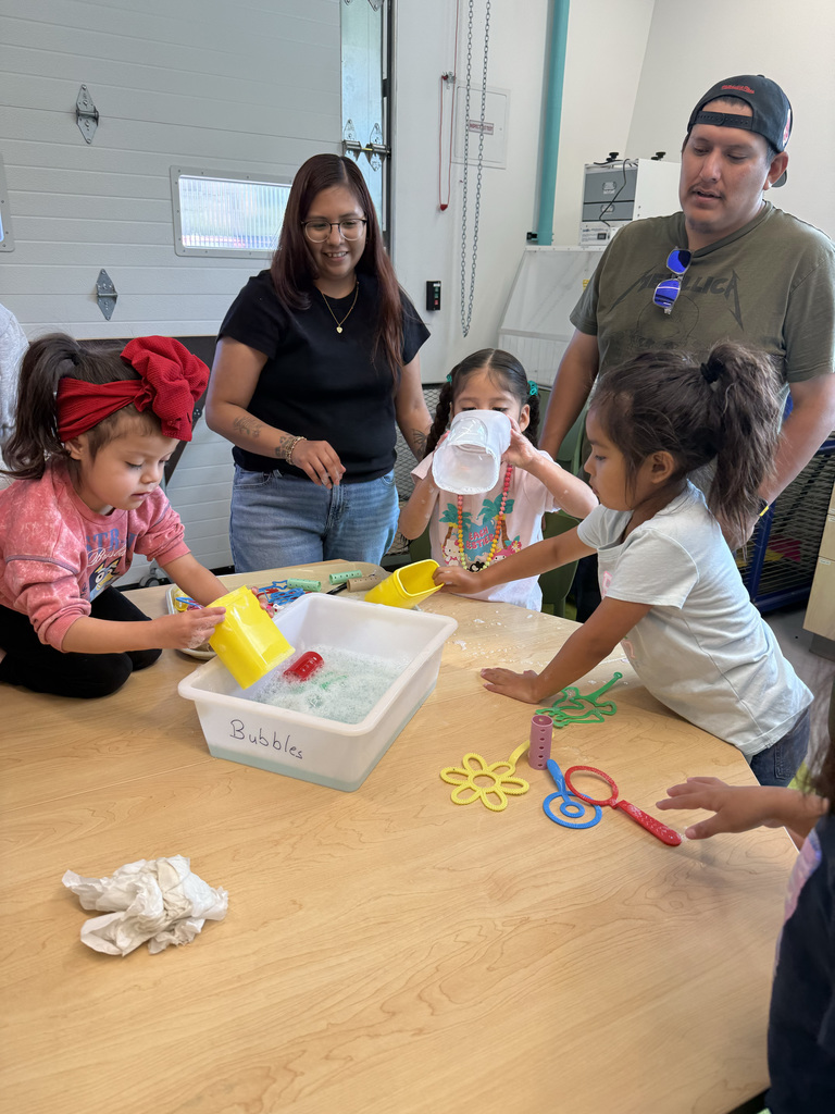 Four children play with toys in a sensory table; a woman in a black shirt and a man in a green shirt watch.