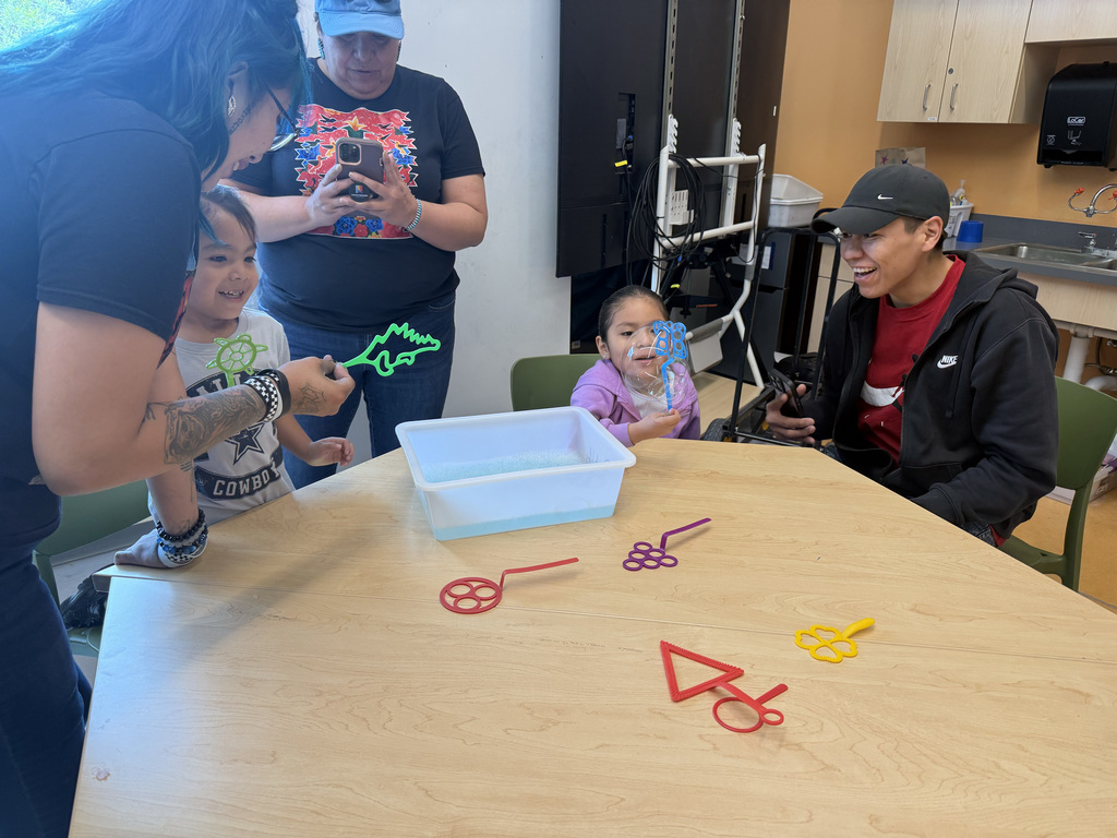People gather around a table with colorful objects. A woman holds a toy. A child sits on a chair.