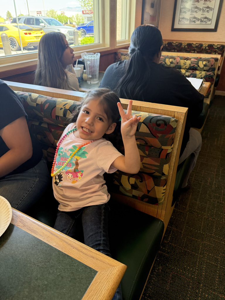 Young girl smiling, making peace sign, seated in a booth at a restaurant, with three adults nearby.