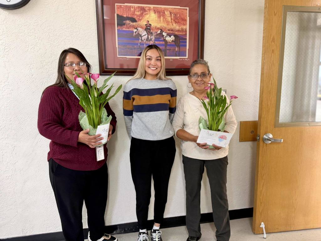 Three women hold tulips in a hallway. A framed picture and an open door are in the background.