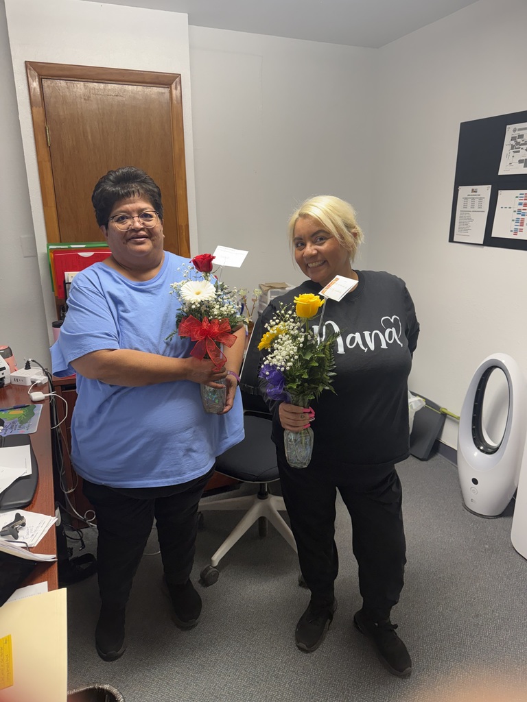 Two women in a room, one holding flowers and the other holding flowers too. Behind them is a wooden door, bulletin board, and a fan.