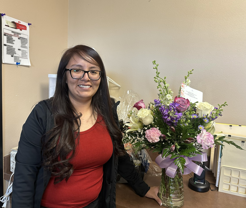 A woman in a red shirt and glasses holds a vase of flowers in a room with beige walls and a bulletin board.