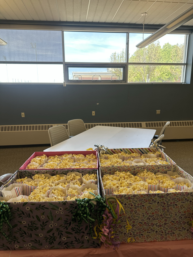 Three boxes filled with popcorn sit on a table. A white table and chairs are behind them. Bright light from a window.