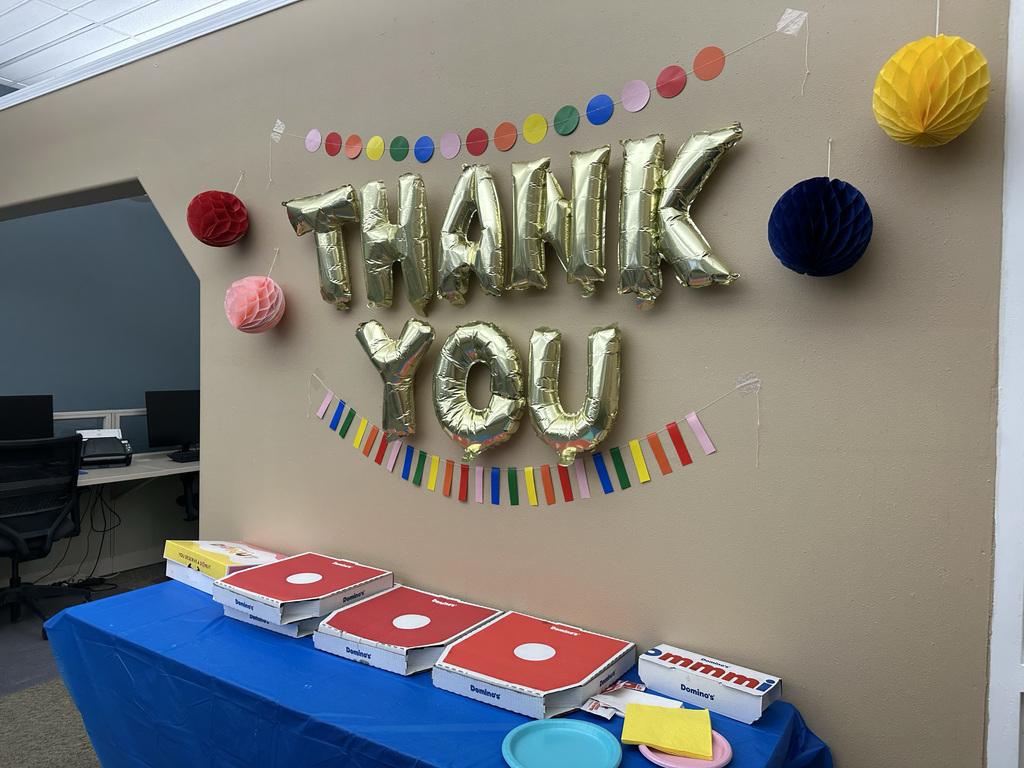 A room displays a "Thank You" banner with colorful balloons and paper garland. A table has pizza boxes and plates.