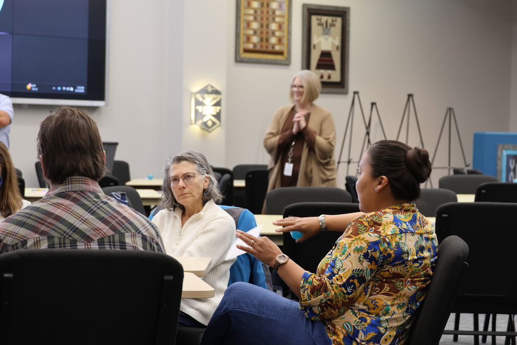 People seated at a table. A woman speaks. A teacher stands near a screen. A framed art hangs on the wall.