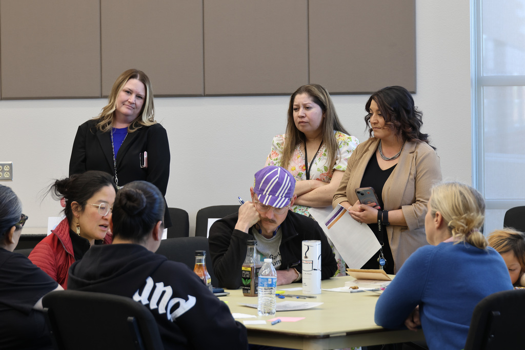 A group of adults, including a man with a purple hat, in a meeting room, seated and standing.