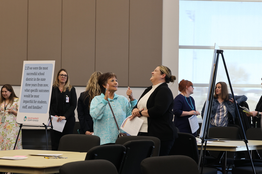 A group of women stand in a meeting room with a table, chairs, and a whiteboard.