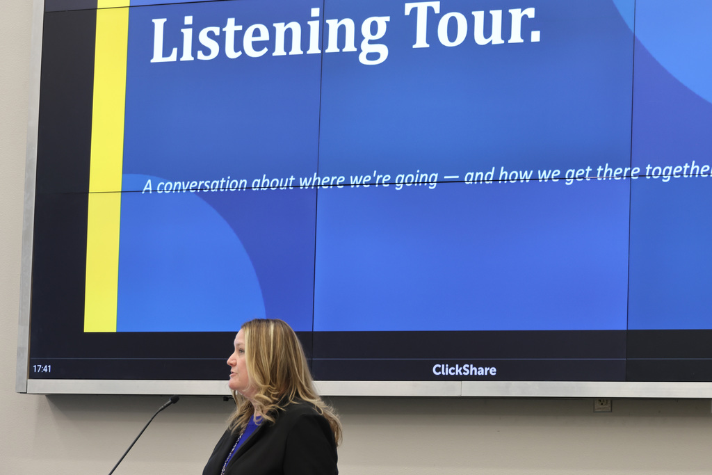 A woman stands at a microphone, facing a large screen with the words "Listening Tour" on a blue background.