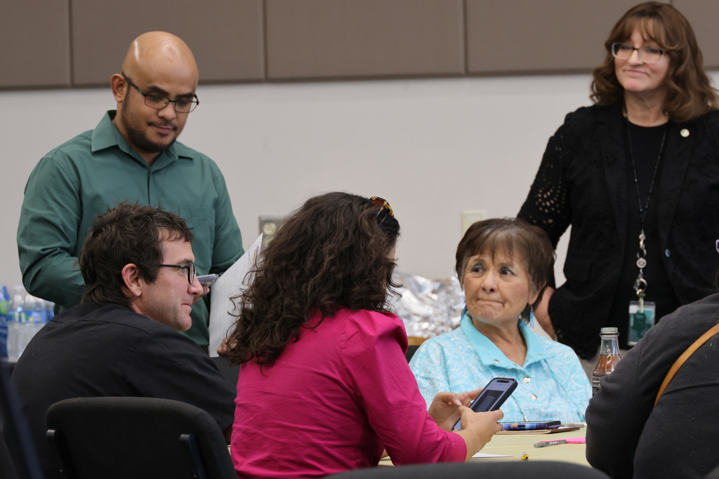 A group of adults sit around a table in a conference room, looking at a cell phone.